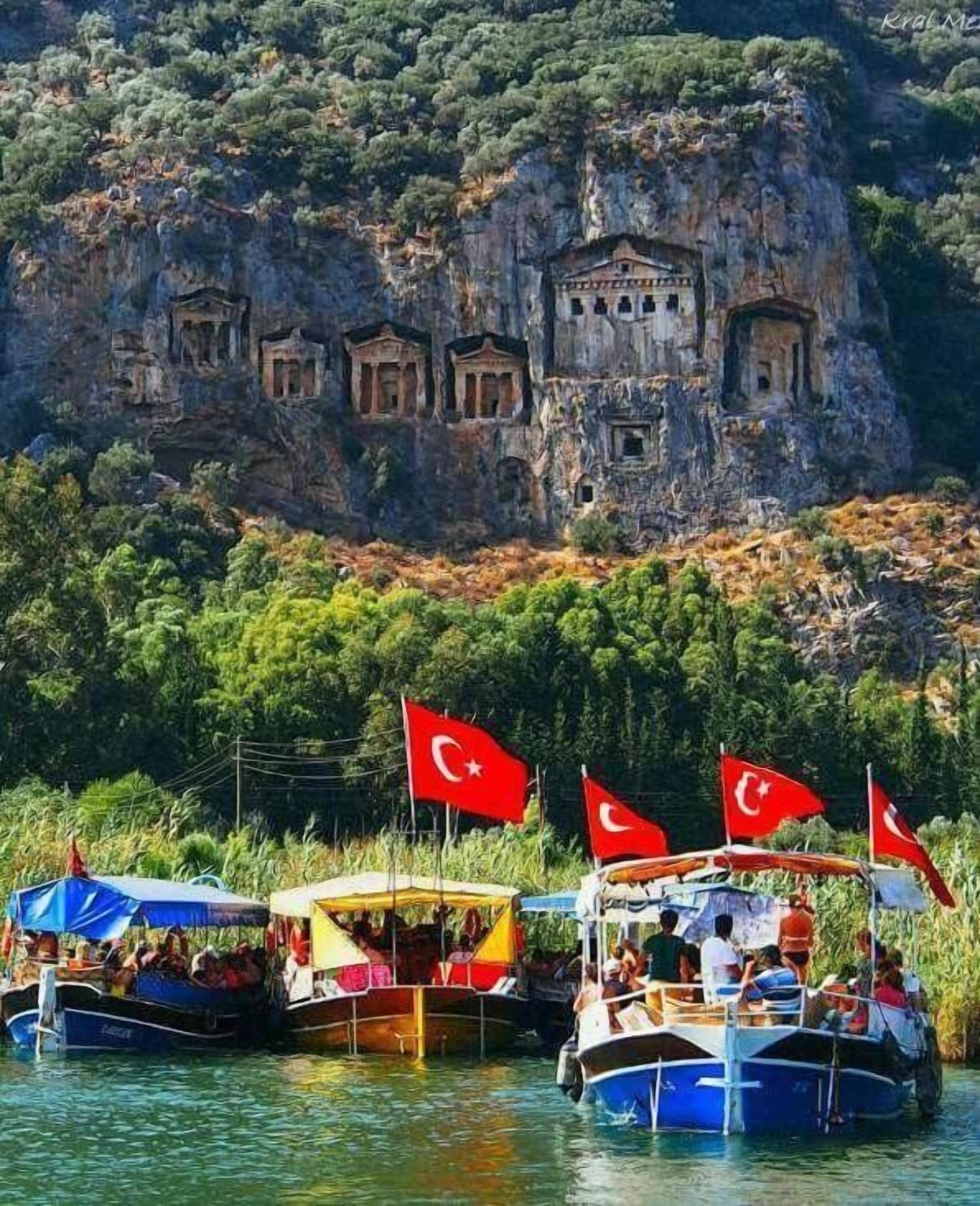 Boats with Turkish flags docked near a rocky cliff with ancient ruins.