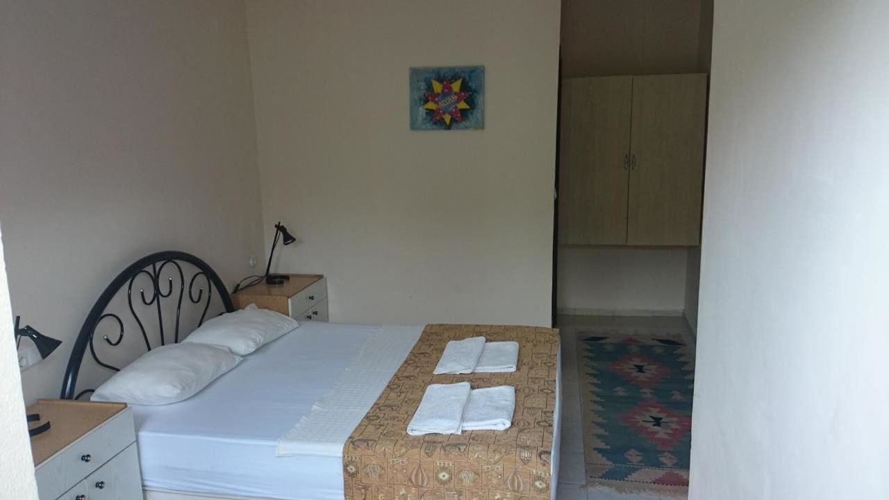 A single bed with a mattress, pillows, and a blanket. There is a small bedside table with a lamp. A colorful artwork hangs on the wall. A wardrobe is visible in the background.