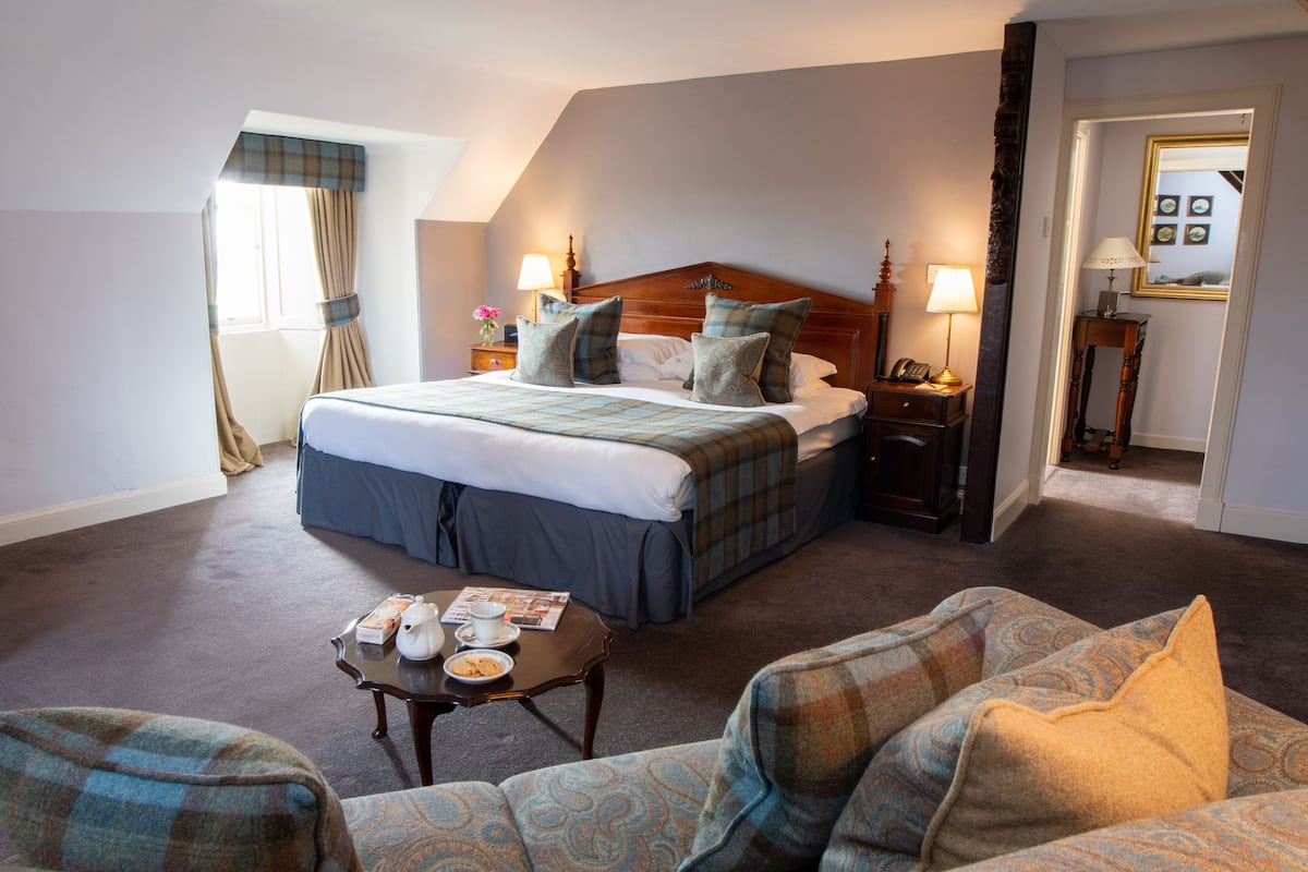 A hotel room with a large bed, a sitting area with a sofa and armchair, a small table with a cup and a book, and a nightstand with a lamp.
