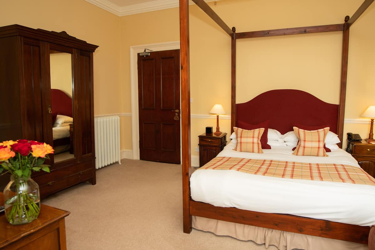 A four-poster bed with a red headboard, a wooden wardrobe, and a vase of flowers on a nightstand.