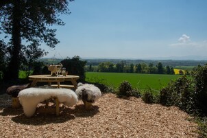 Una mesa de madera con una botella y vasos, rodeada de taburetes blancos y mullidos, sobre una superficie de grava con vistas a un campo verde y colinas distantes.