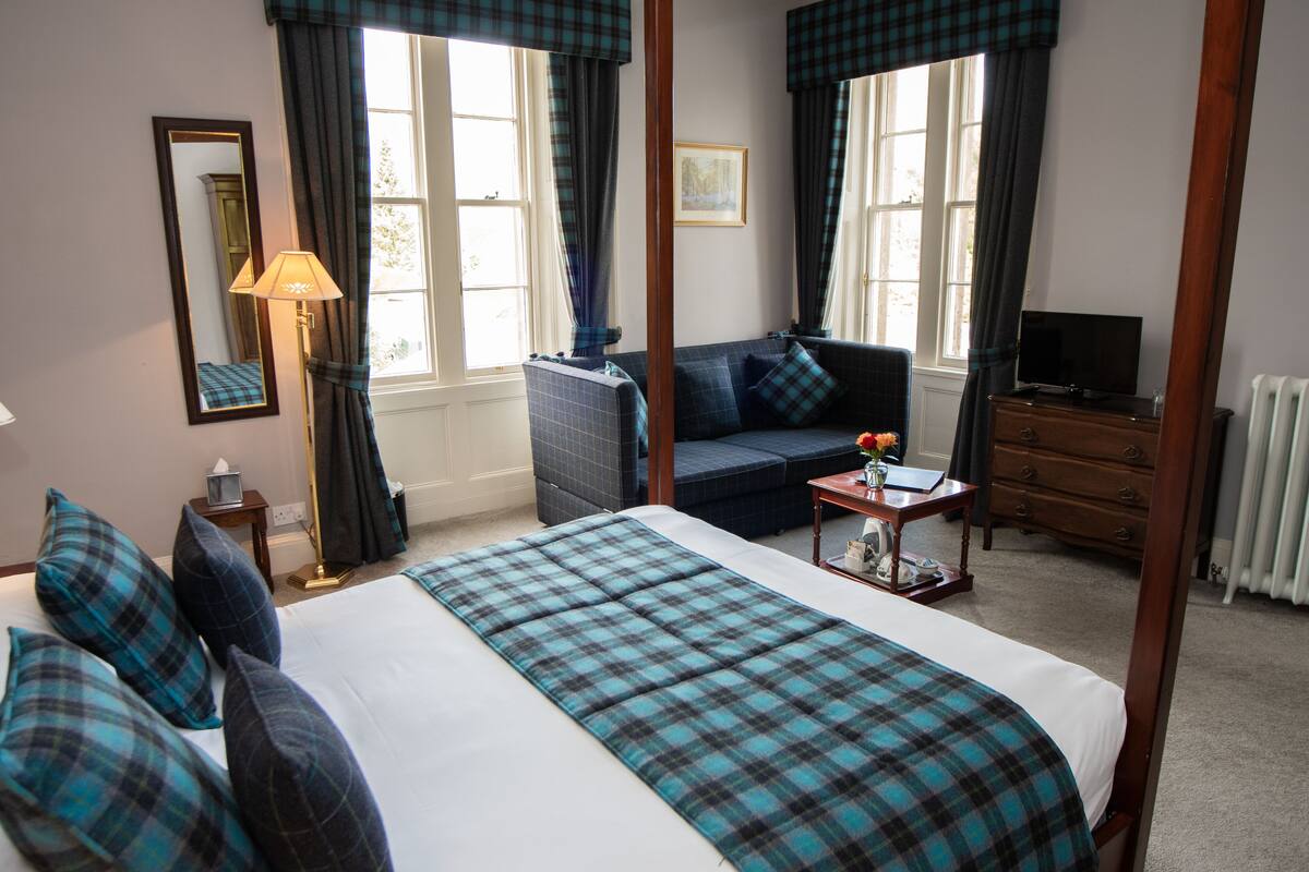 A four-poster bed with a plaid bedspread, a sofa, a television, and a dresser in a well-lit room with large windows and curtains.