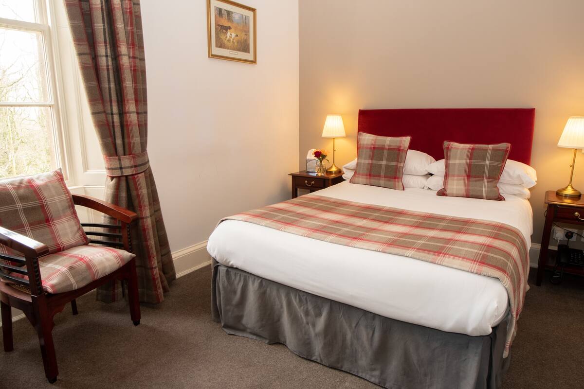 A neatly arranged bedroom with a large bed, a chair, a nightstand with a lamp, and a window with curtains.
