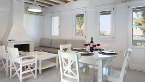 Un espace repas moderne, avec des meubles blancs, une cheminée et de grandes fenêtres donnant sur l’extérieur.