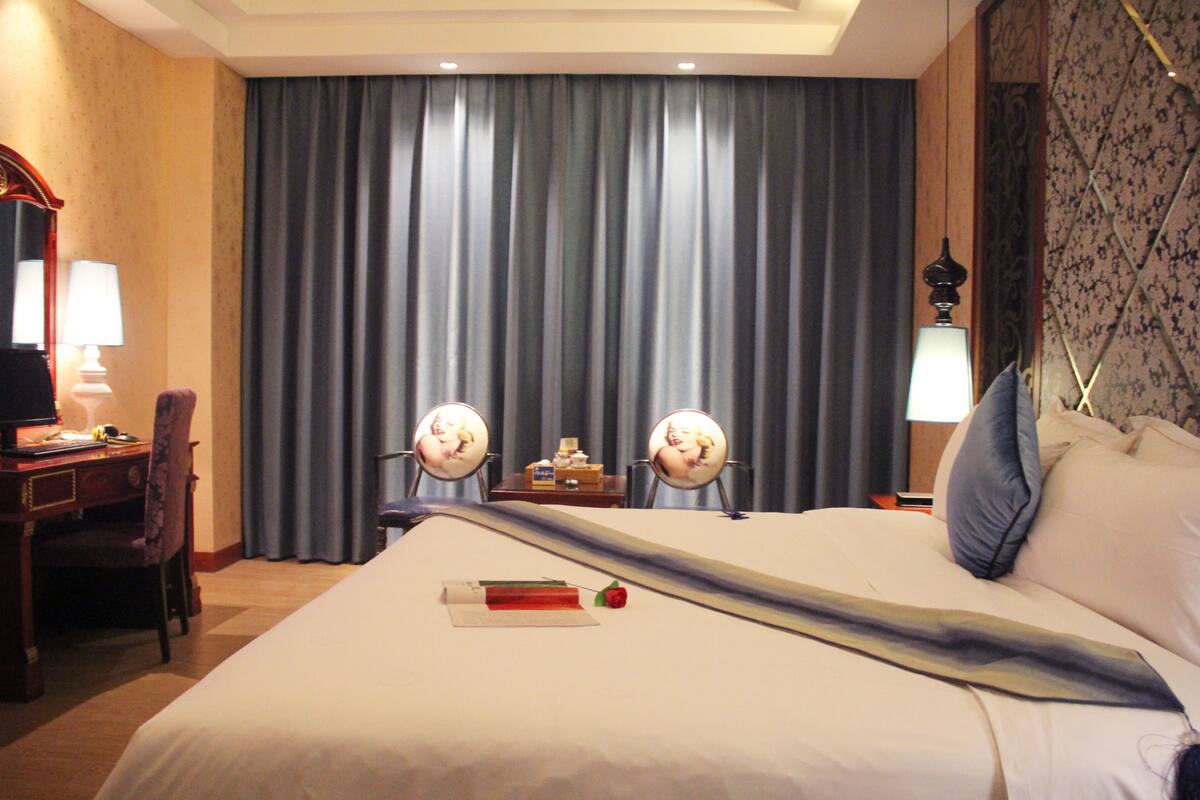 Una habitación de hotel con una cama grande, dos mesitas de noche con lámparas, un escritorio con una silla y un ventanal con cortinas.