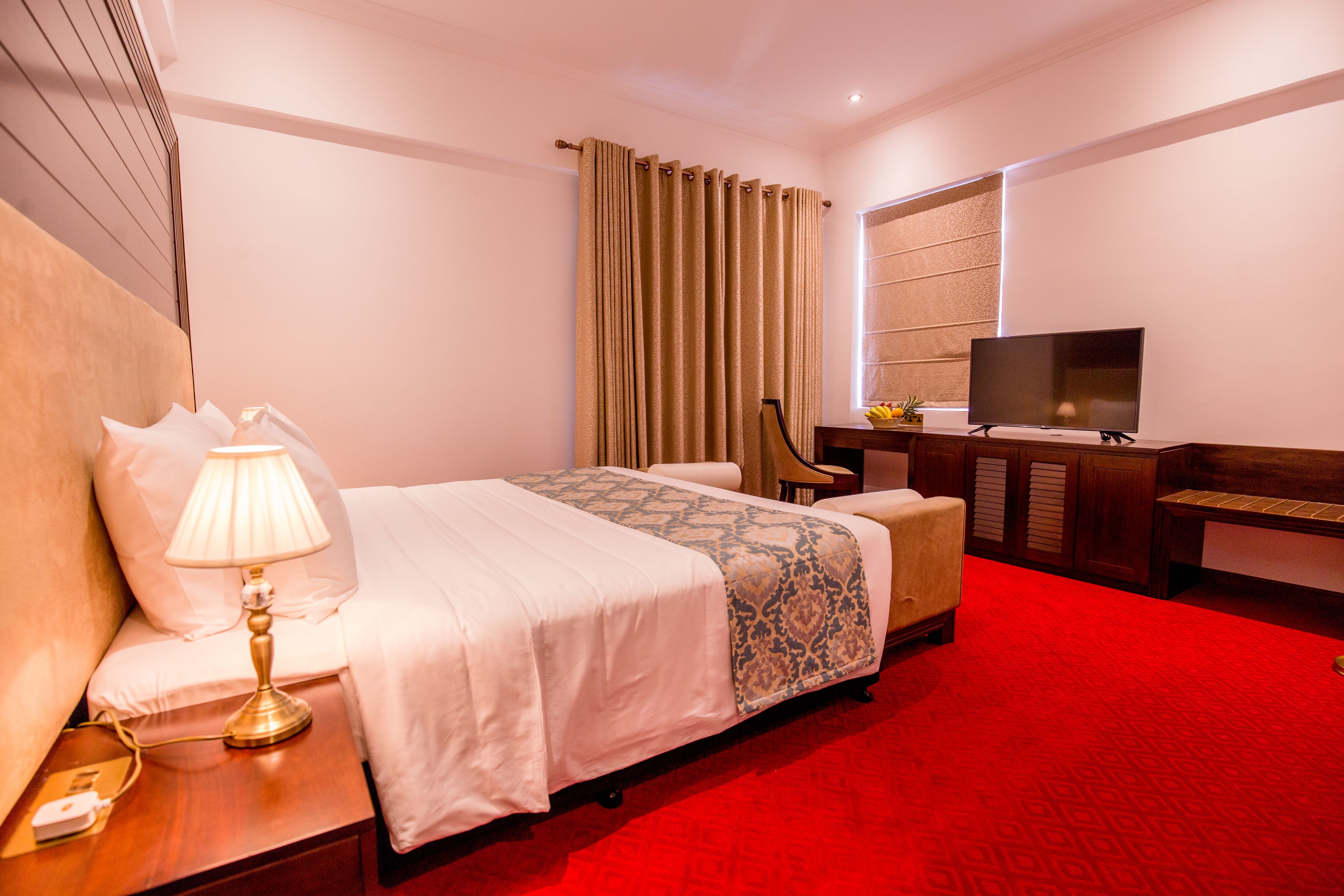 A hotel room with a bed, a desk with a lamp, a television, and a red carpet.