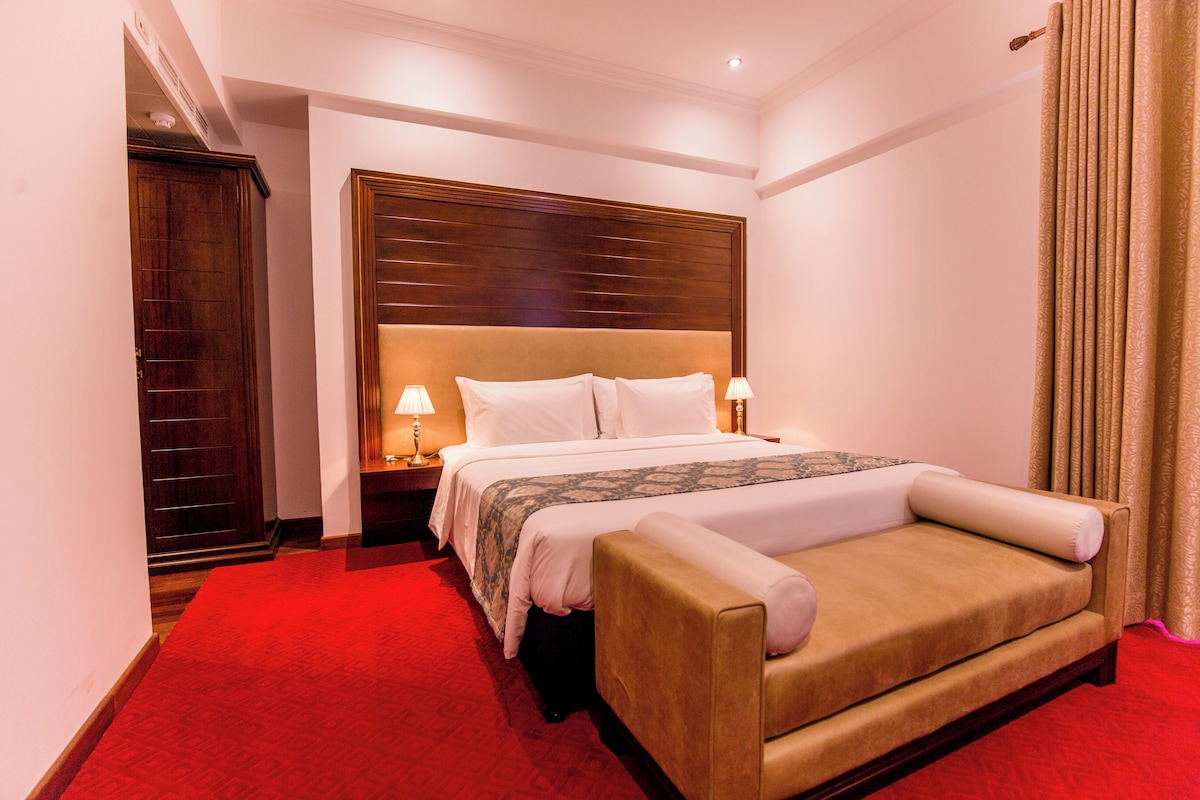 A hotel room with a bed, a nightstand, a lamp, and a red carpet.