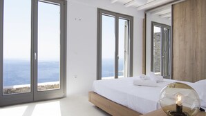 Une chambre avec un grand lit, une table de chevet en bois et une vue sur la mer grâce à de grandes fenêtres.