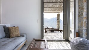 Un salon moderne avec un canapé gris, une table basse en bois et un pouf à motifs. Une porte-fenêtre coulissante donne accès à une terrasse extérieure avec une table et des chaises en bois.