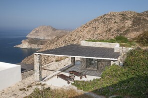 A coastal building with a stone exterior, a flat roof, and a patio area with two wooden tables and chairs. - Serifos Houses Kalo Ampeli (Serifos)