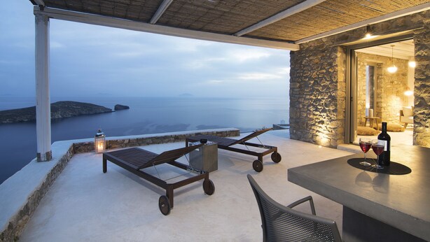 Une terrasse avec vue sur la mer, un mur en pierre, une table avec une bouteille et des verres, et deux fauteuils de jardin.