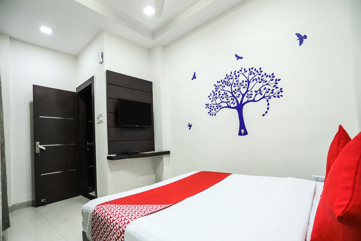A hotel room with a bed, a TV mounted on the wall, a red and white bedspread, and a wall decal of a tree with birds.