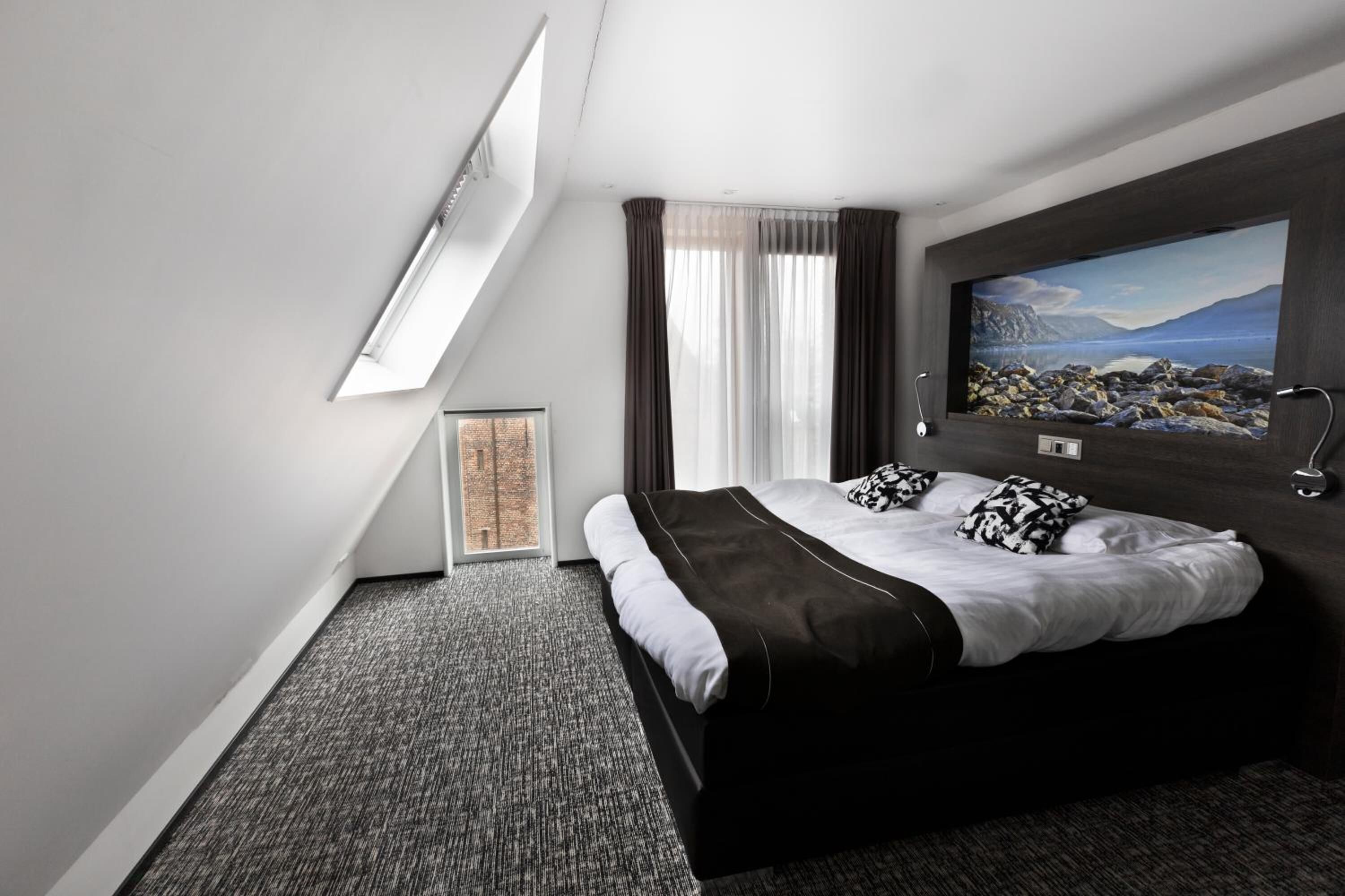 A modern hotel room with a large bed, a painting on the wall, and a skylight.