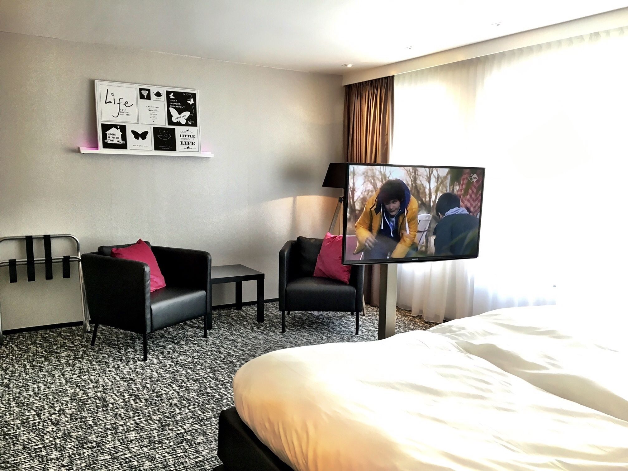 Ein Hotelzimmer mit einem Bett, zwei schwarzen Sesseln, einem kleinen Tisch, einem Flachbildfernseher und einer Wand mit motivierenden Postern.