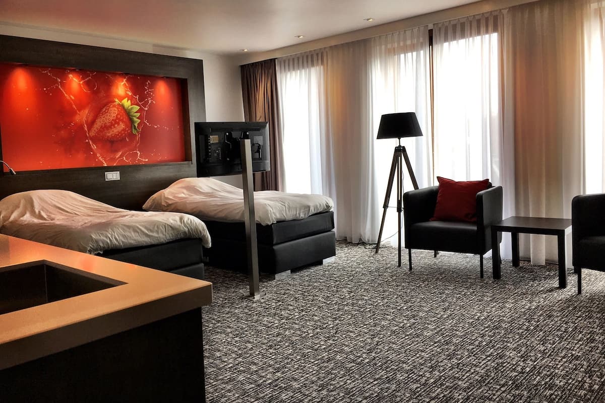 A hotel room with two beds, a desk, a chair, a floor lamp, and a large window with curtains.