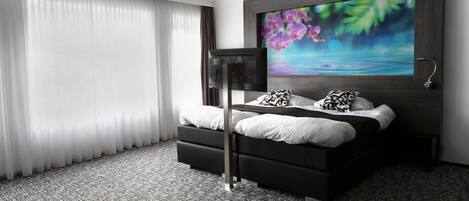 Una habitación de hotel moderna con una cama grande, un televisor de pantalla plana y un ventanal con cortinas translúcidas.