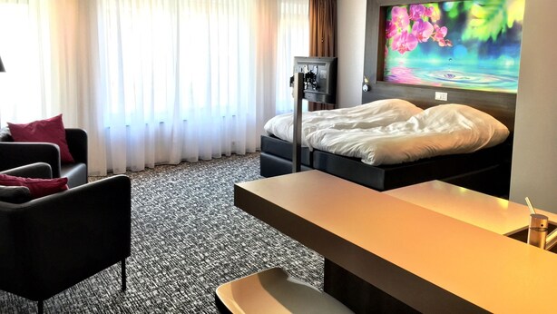 A hotel room with a bed, a desk, a chair, a TV, and a large window with curtains. - Apart Food and Drinks (Zwolle)