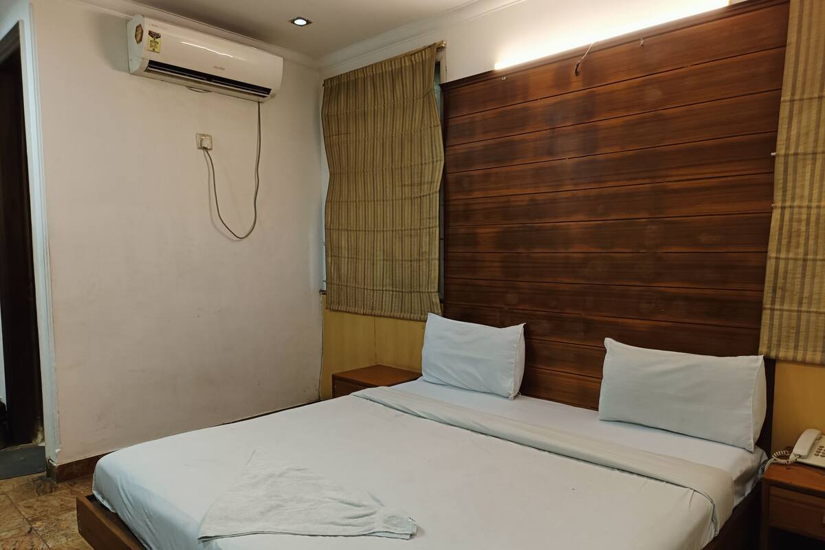 A hotel room with a wooden headboard, a bed with white linens, a bedside table with a phone, and an air conditioning unit on the wall.