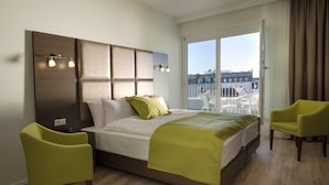 Ein modernes Hotelzimmer mit einem großen Bett, einem Balkon mit Blick auf Gebäude und zwei grünen Stühlen.