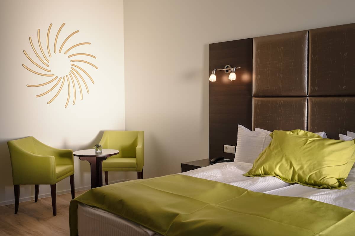 Ein Hotelzimmer mit einem Bett, zwei grünen Stühlen, einem kleinen Tisch, einem Wandbild und einer Nachttischlampe.