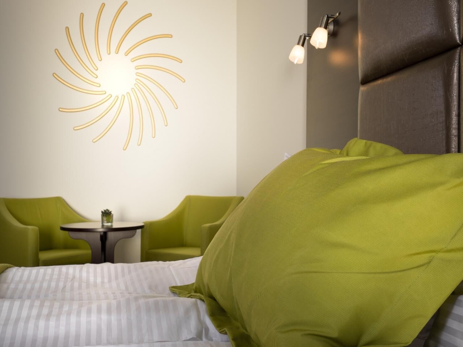 A hotel room with a bed, two green armchairs, a small round table, and a wall-mounted light fixture.