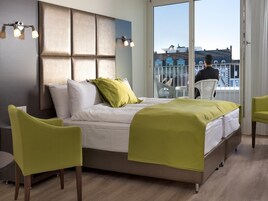 Quarto de hotel moderno com uma cama grande, cadeiras verdes e vista para a cidade.