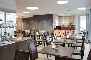 Um restaurante moderno com várias mesas e cadeiras, um balcão exibindo pratos e uma parede decorativa com motivos de folhas de palmeira.