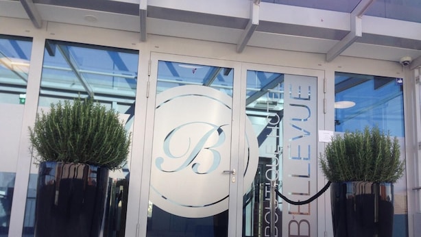 Ein Hotel mit Eingangsbereich und Glastür. Auf der Tür befindet sich ein großes 'B' Logo und der Name 'Bellevue'. Auf beiden Seiten der Tür stehen je ein Blumentopf.