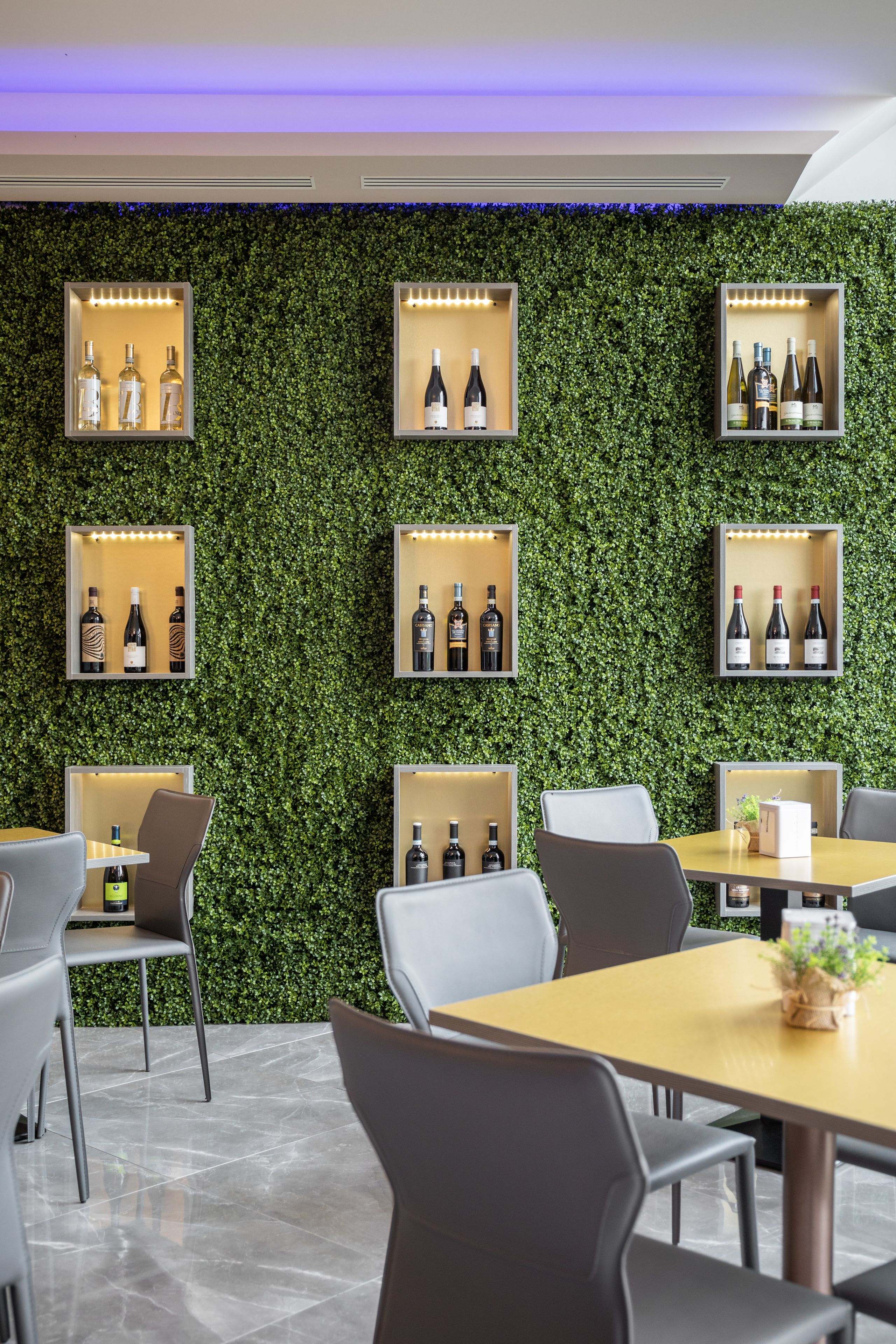 A modern interior with a green wall and shelves displaying bottles. There are tables and chairs arranged for dining.