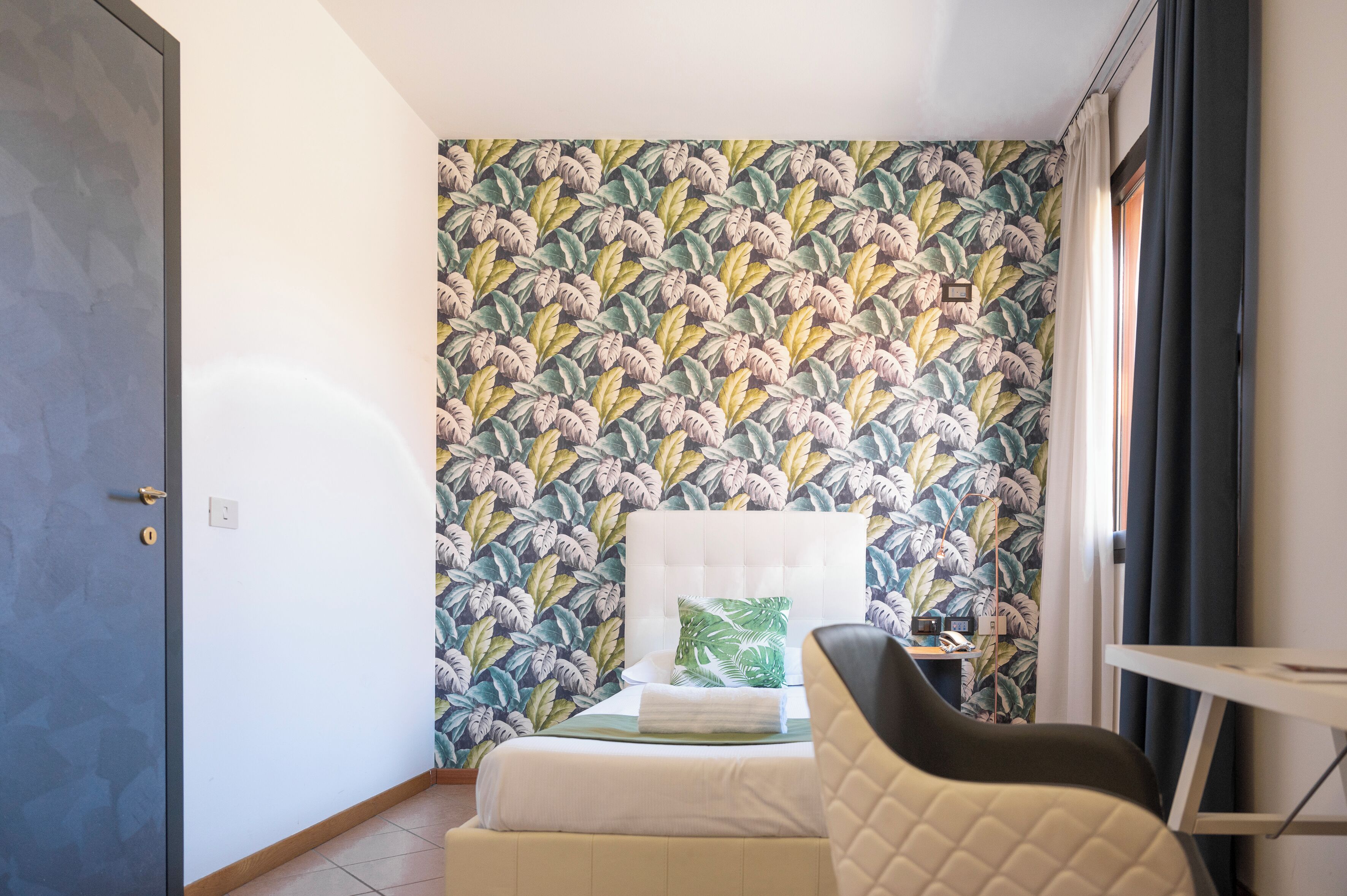 Una habitación pequeña con cama, una silla y una mesa. Presenta un papel tapiz estampado y una puerta.
