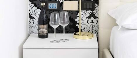 Una mesita de noche blanca con una botella de vino y dos copas, un objeto decorativo pequeño y un enchufe.