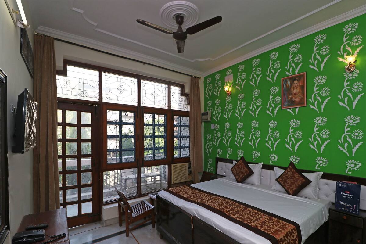 Habitación de hotel con cama, escritorio con silla, ventilador de techo y ventana grande con vistas al exterior.
