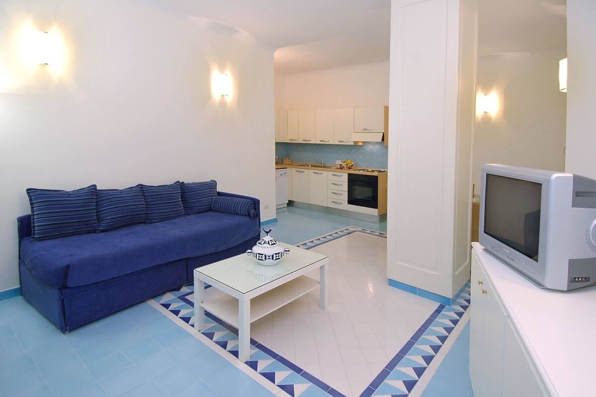 A compact living space with a blue sofa, a small white table with a teapot, a television, and a kitchen area in the background.