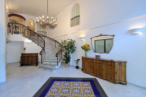 A spacious foyer with a marble staircase, a chandelier, a wooden console, and a patterned rug. - Aurora Residence (Amalfi)