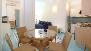 Un espacio habitable compacto con una mesa redonda de vidrio rodeada de sillas de mimbre, un sofá azul con cojines, una kitchenette con estufa y fregadero, y un pequeño armario blanco.