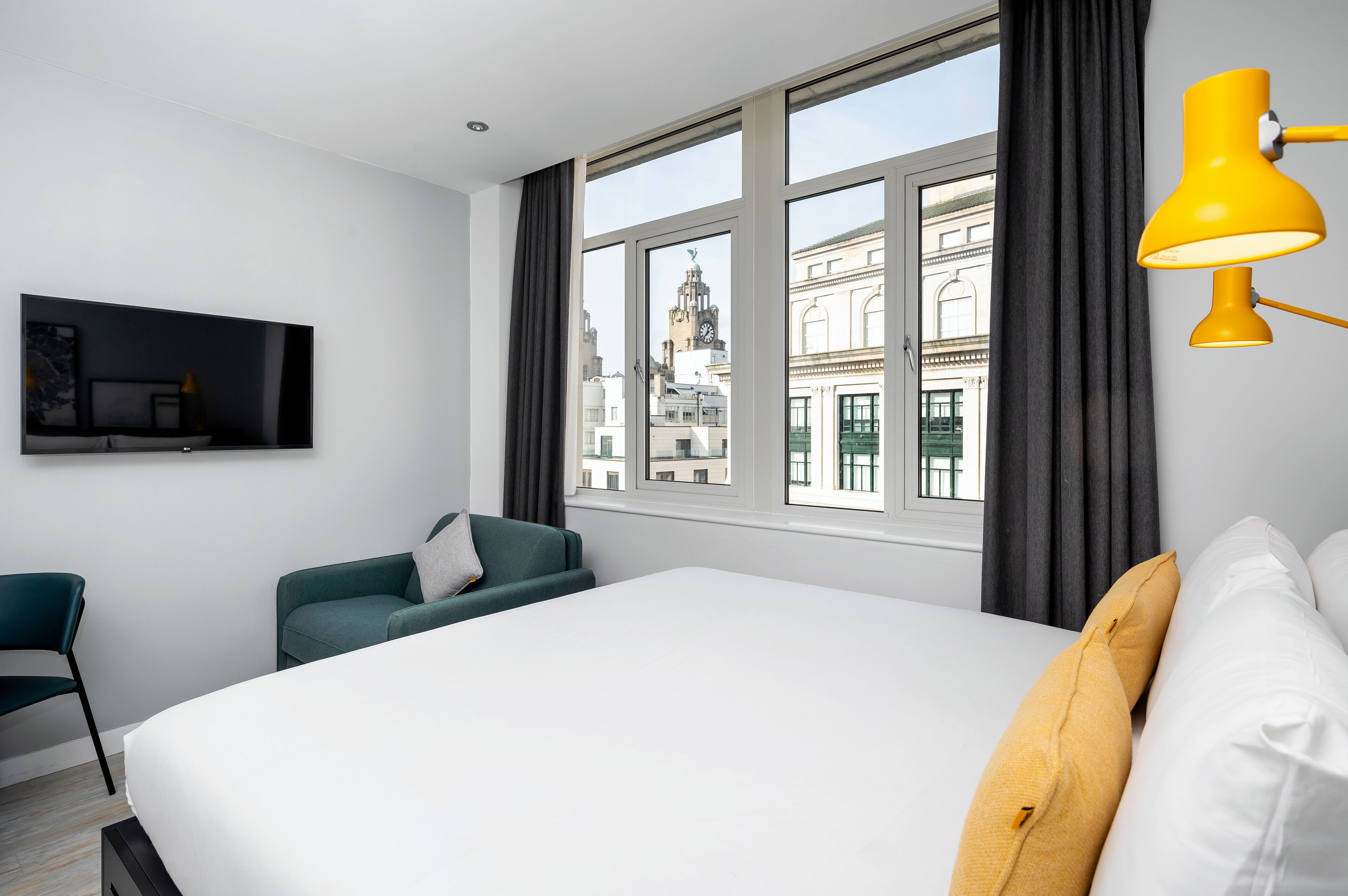 Camera d'albergo moderna con un letto grande, una poltrona verde, una TV a schermo piatto e vista sugli edifici della città attraverso ampie finestre.