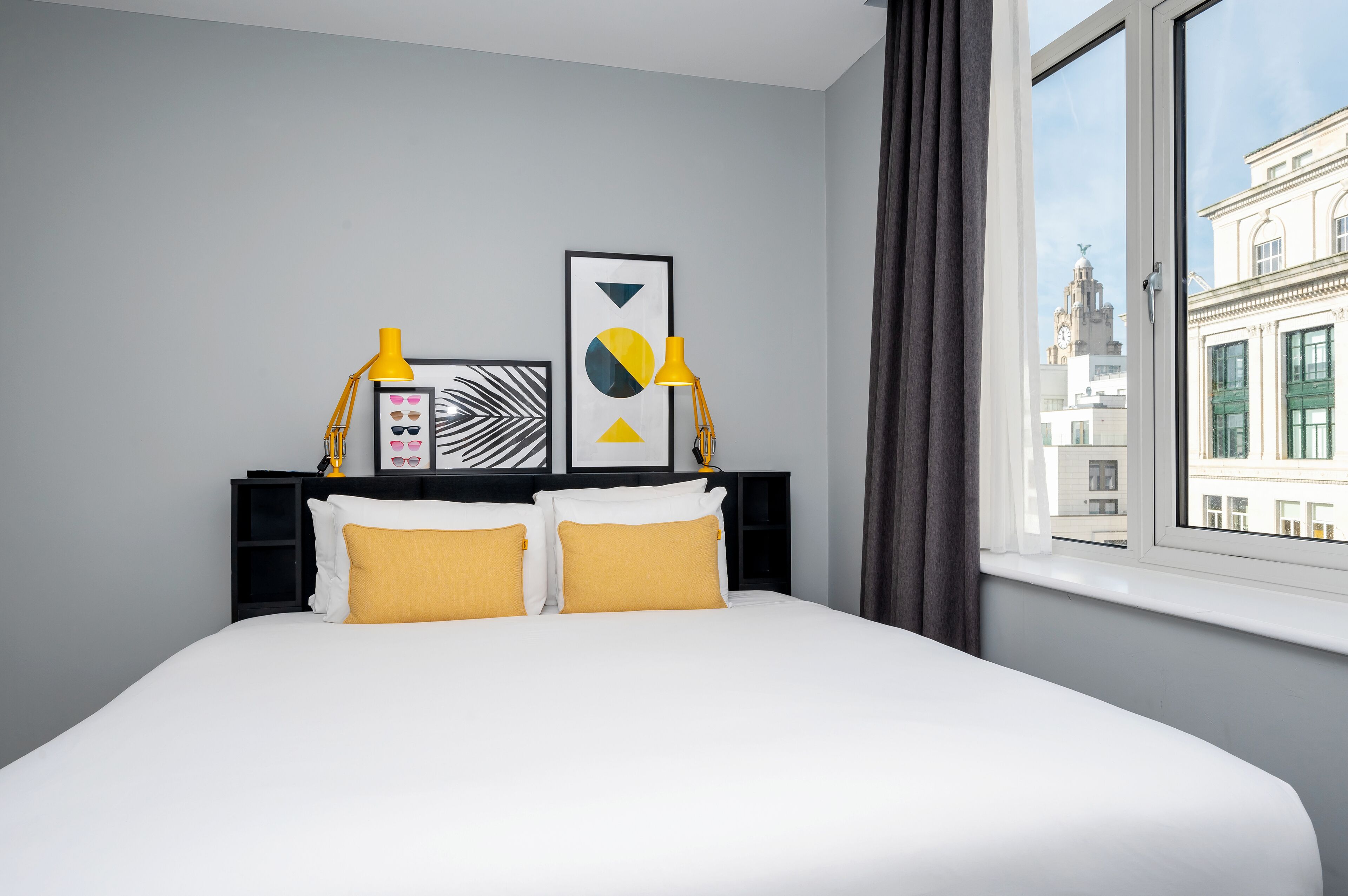 A hotel room with a bed, two yellow pillows, a black headboard, a framed picture, and a window with a view of a building.
