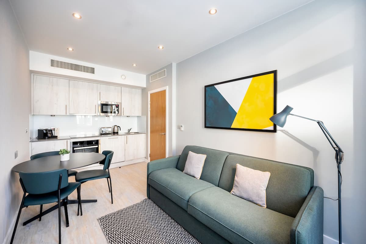 A modern kitchen with white cabinets, a dining table with chairs, and a grey sofa with a lamp.