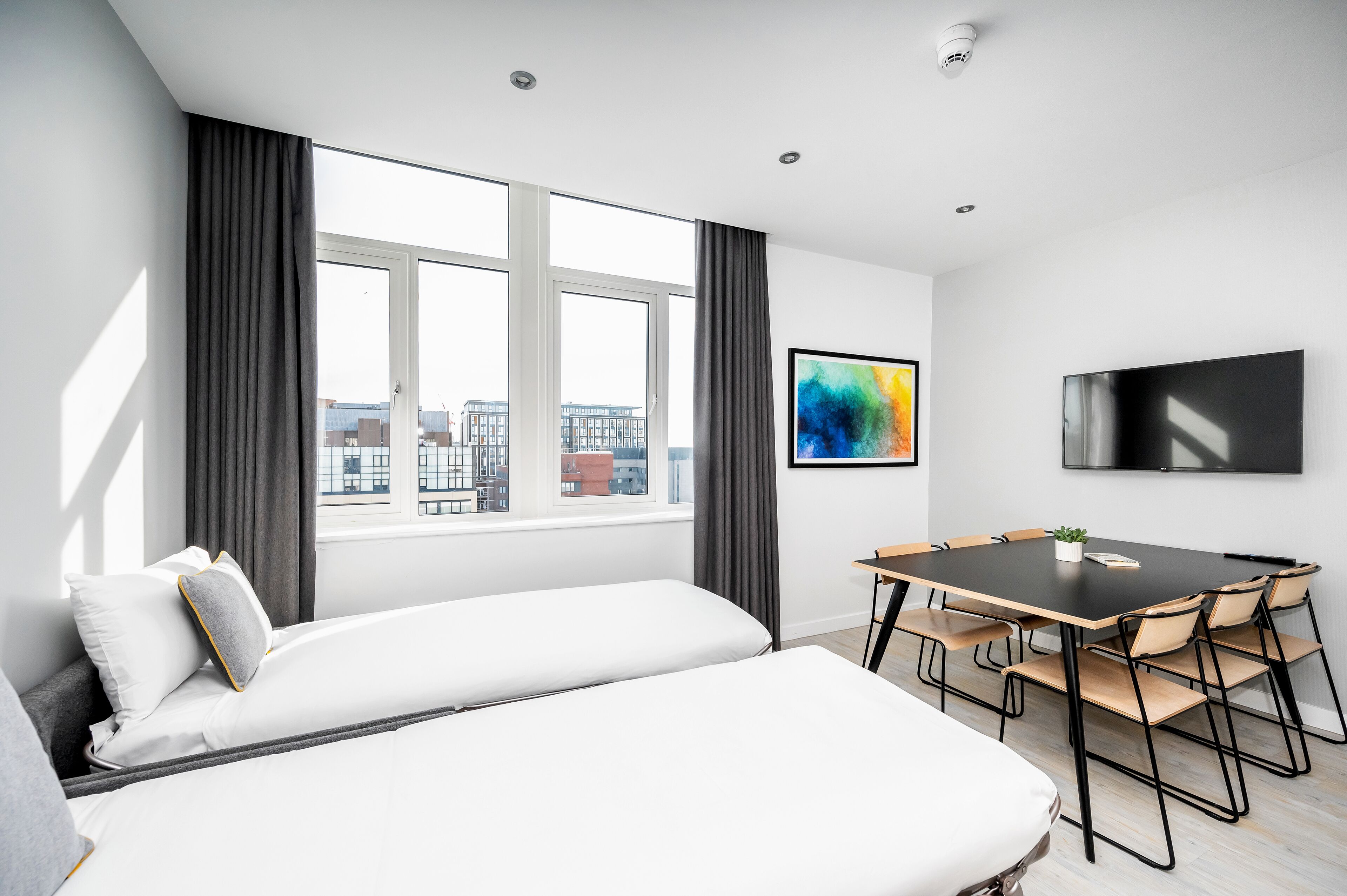 Ein modernes Hotelzimmer mit einem großen Bett, einem Esstisch mit Stühlen, einem Flachbildfernseher und Blick auf die Stadt.