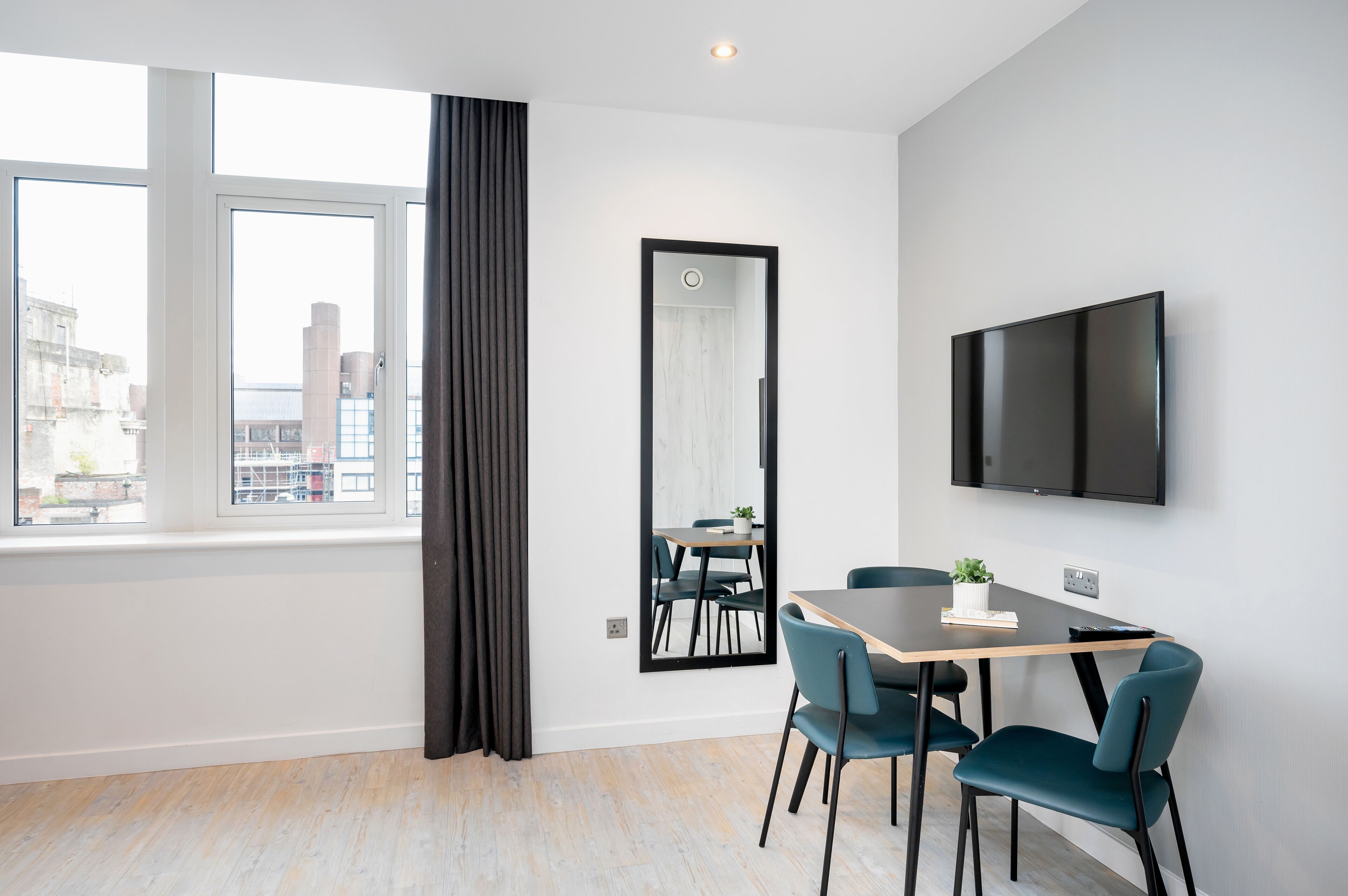 A modern room with a large window, a flat-screen TV mounted on the wall, a dining table with chairs, and a mirror.