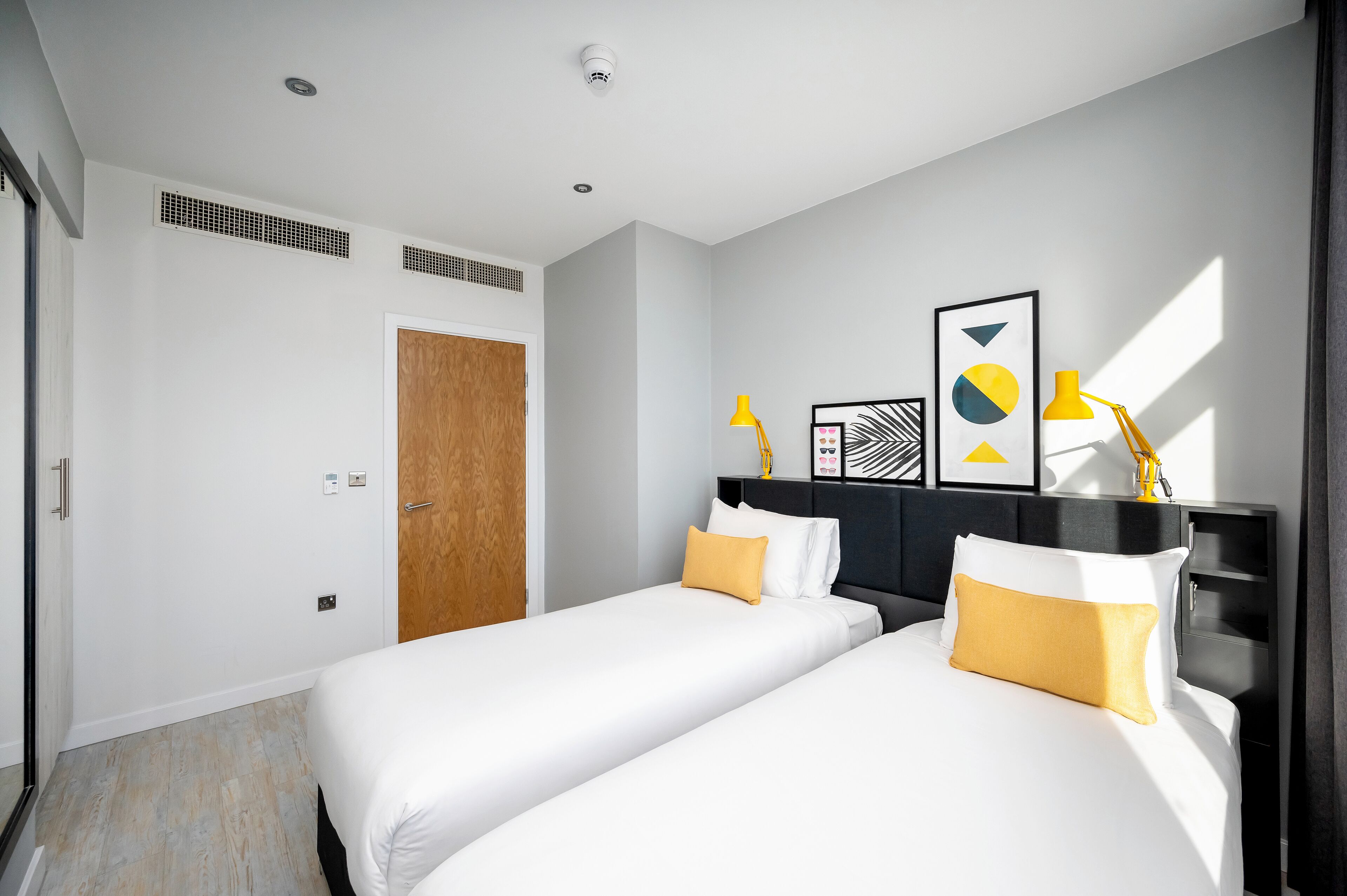 Habitación de hotel con dos camas, una puerta de madera y un diseño moderno, con cabecero negro y lámparas amarillas.