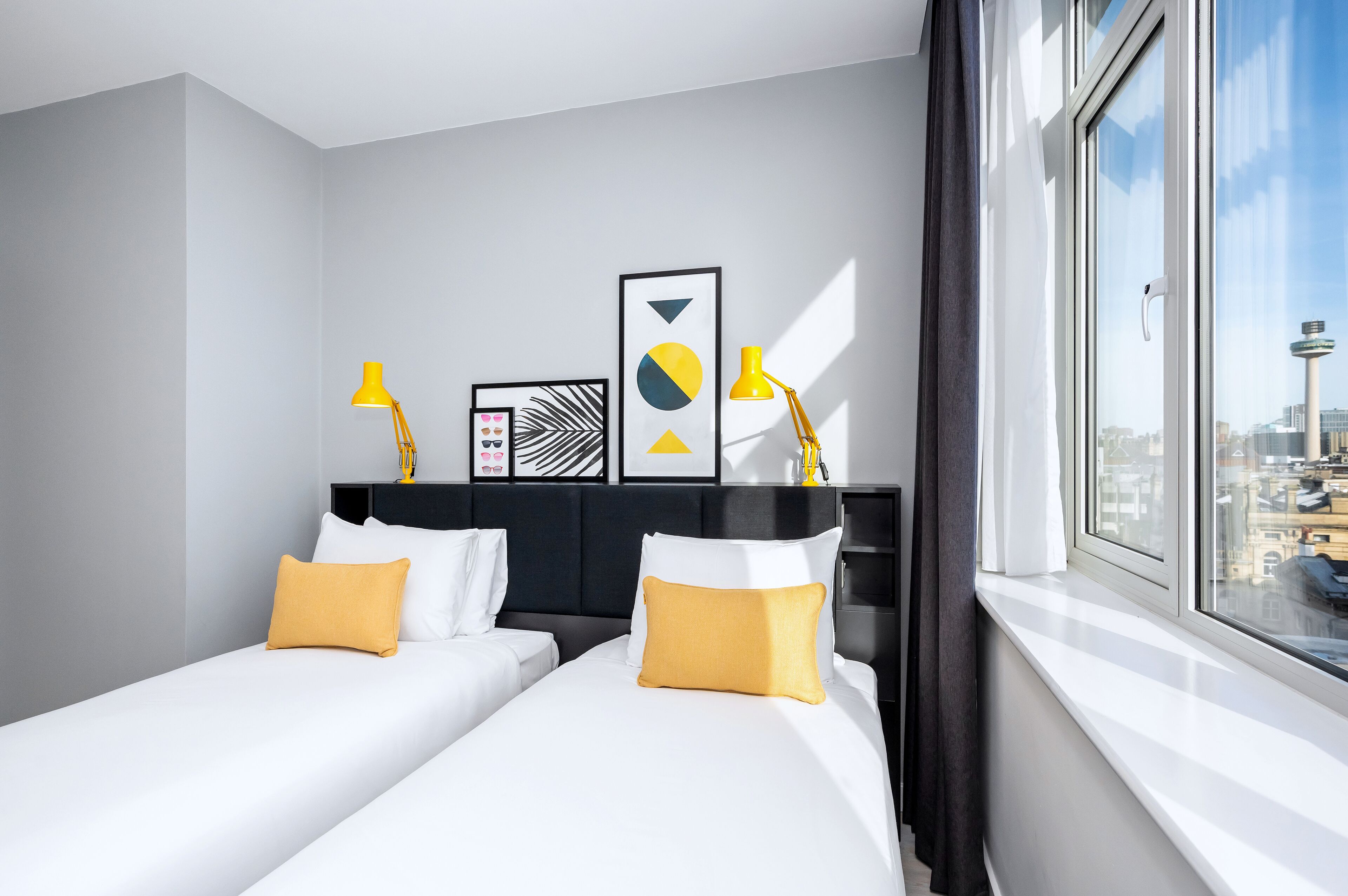 A modern hotel room with two beds, a large window with a city view, and decorative wall art.