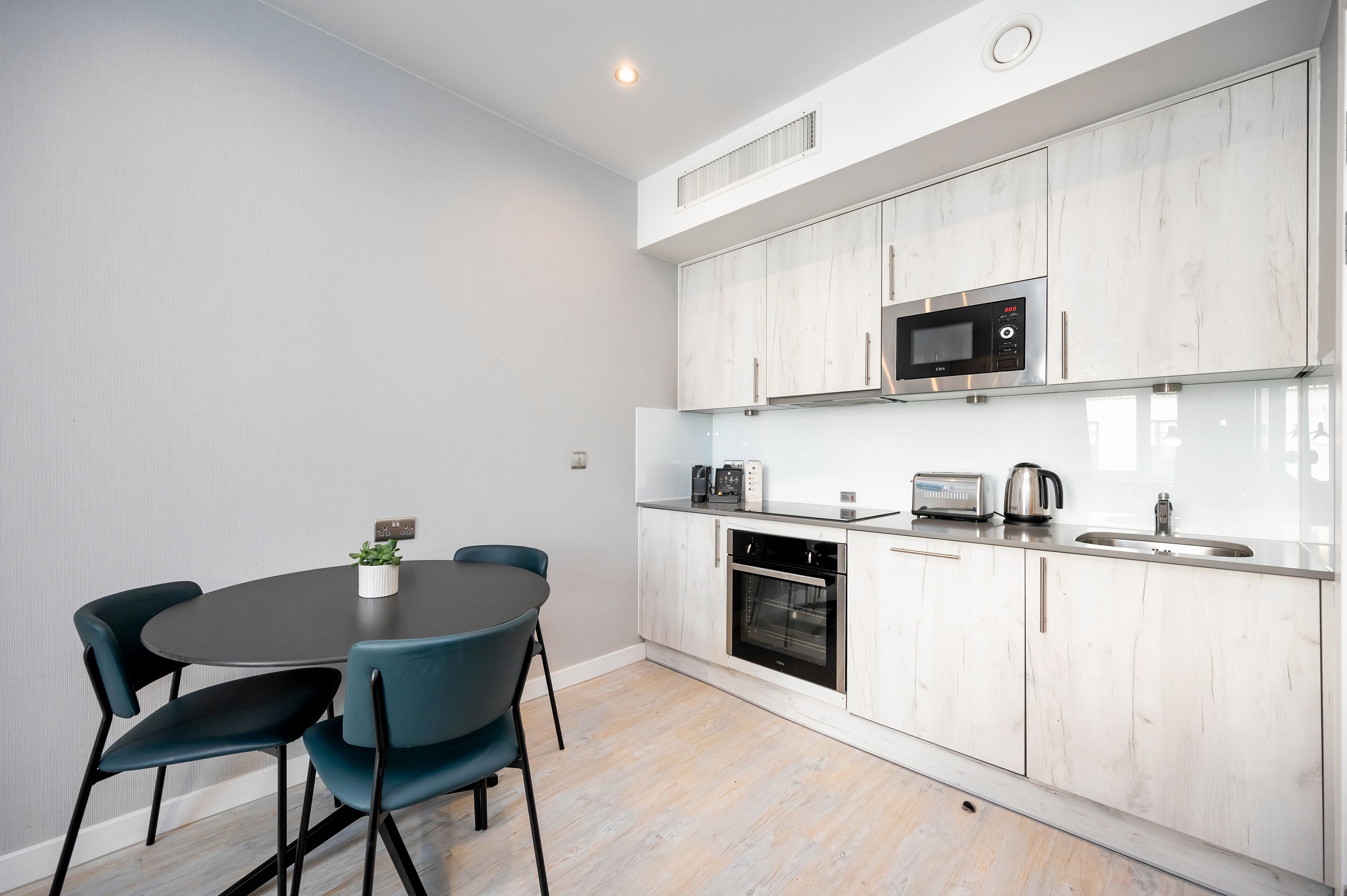 Cocina moderna con armarios blancos, microondas, tostadora y fregadero. Hay una mesa redonda con sillas y una planta en maceta.