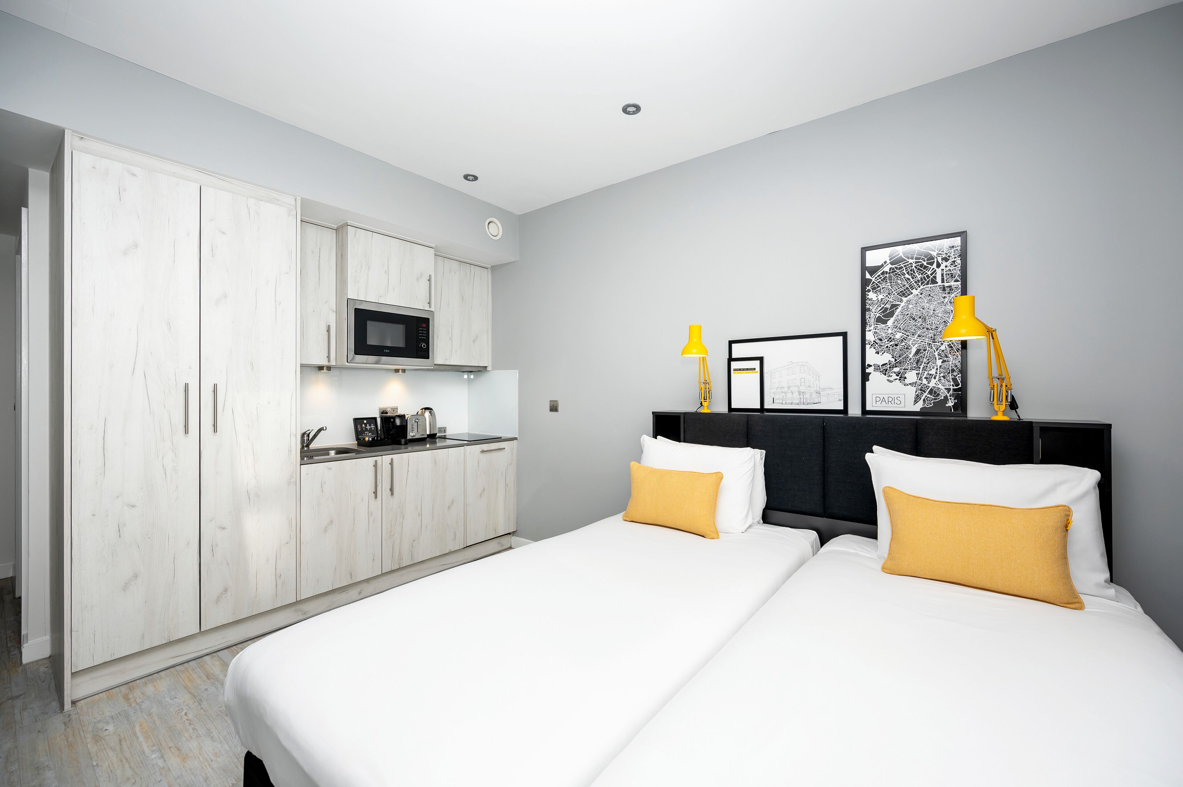 A modern hotel room with a bed, white wardrobe, kitchenette, and a framed artwork on the wall.