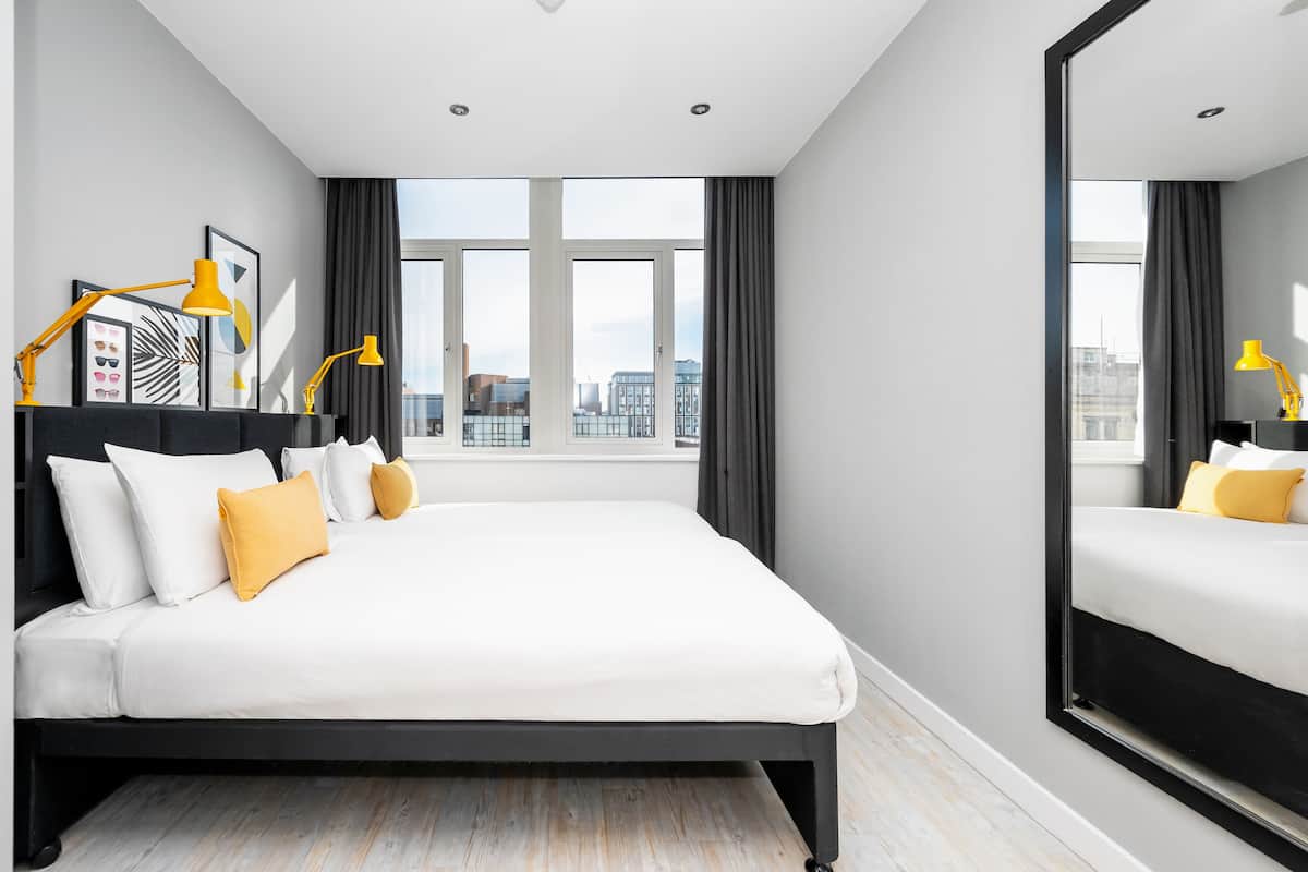 A modern hotel room with a large bed, a city view, and a mirror reflecting the room.