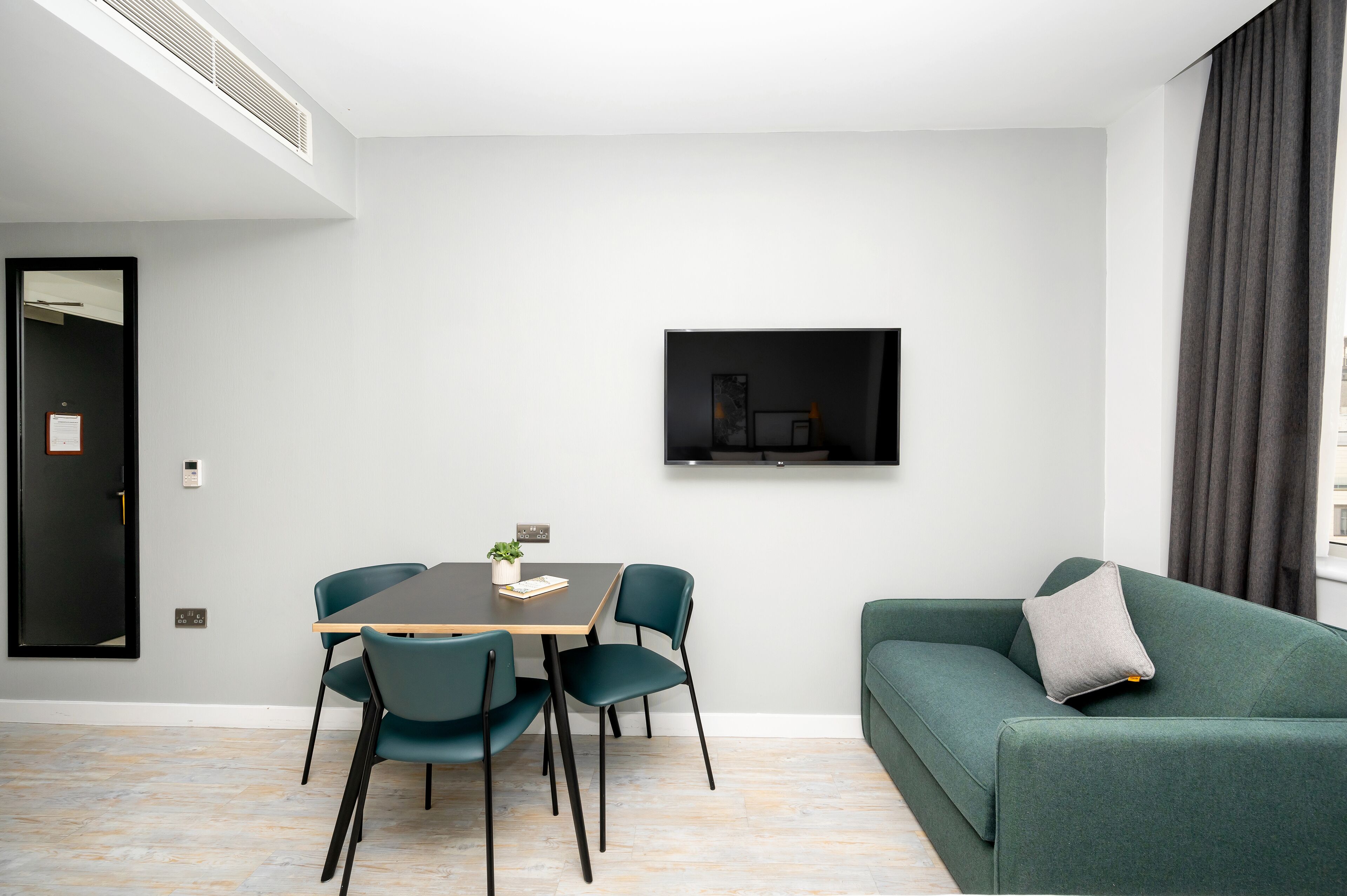A modern living room with a dining area, a flat-screen TV mounted on the wall, and a green sofa with a pillow.