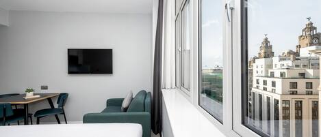 A modern hotel room with a large window, a flat-screen TV mounted on the wall, a dining table with chairs, and a green sofa.