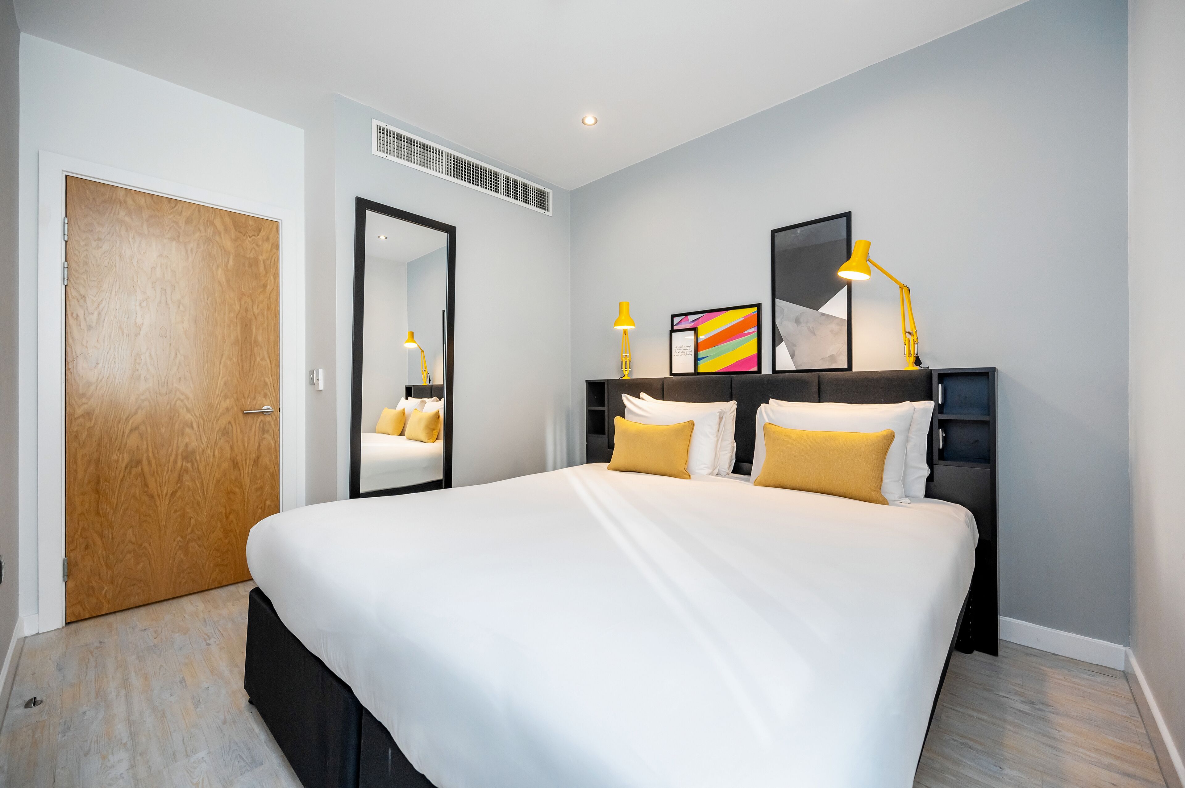 Una habitación de hotel moderna con una cama grande, una mesita de noche con lámpara, un espejo y un cuadro enmarcado en la pared.
