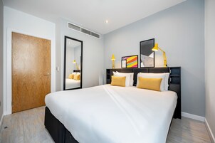 A modern hotel room with a large bed, a bedside table with a lamp, a mirror, and a framed picture on the wall.