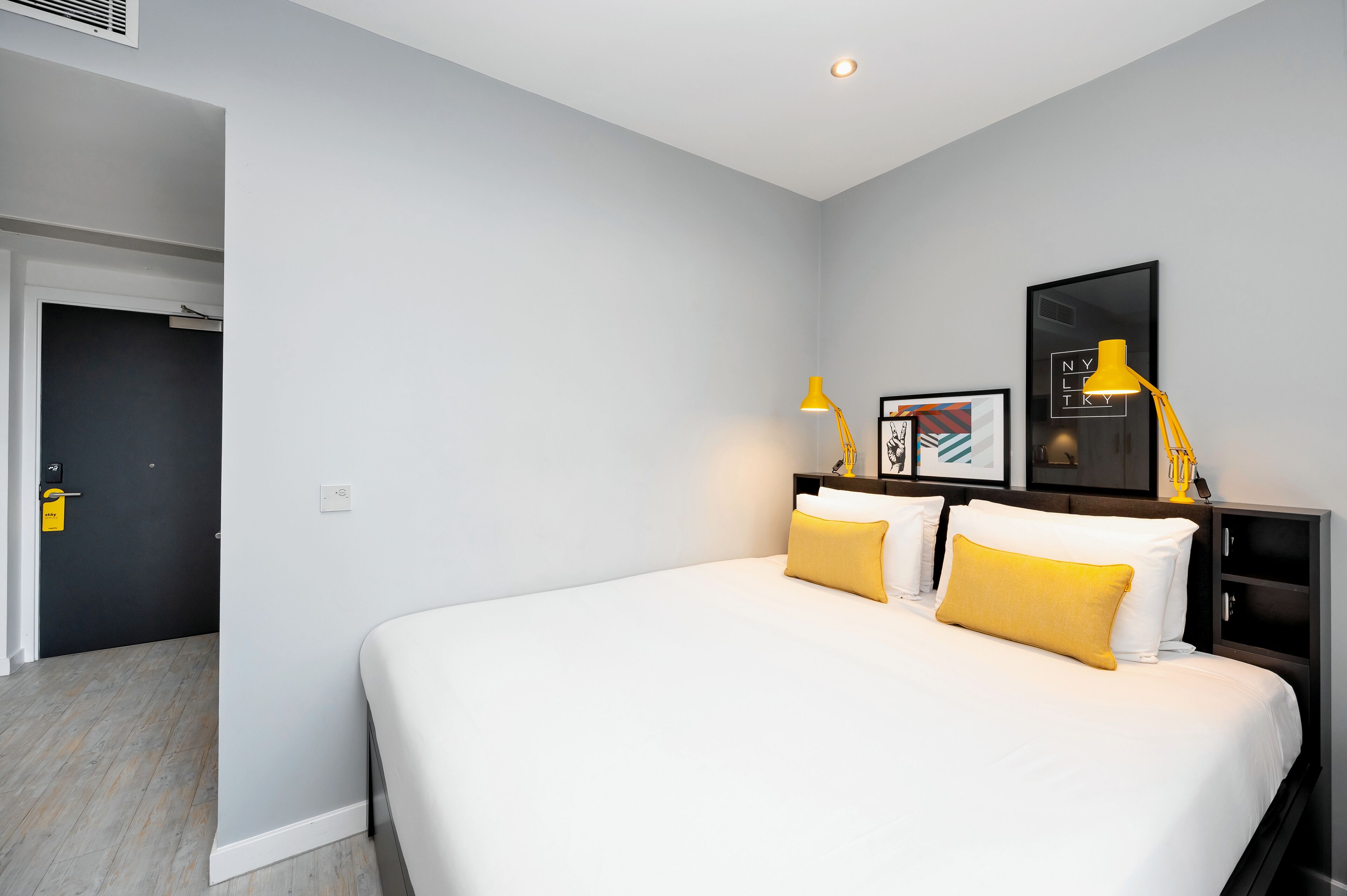 Una habitación de hotel moderna con una cama grande, una mesita de noche, un cuadro enmarcado y una lámpara amarilla.